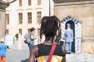 PRAGUE - AUGUST 29: Black woman in selective focus from behind with dreadlocks lifts mobile to yake photo of Guards Prague Castle Guard in blue uniform at attention while other tourists walk around looking at Prague Castle and the guards
