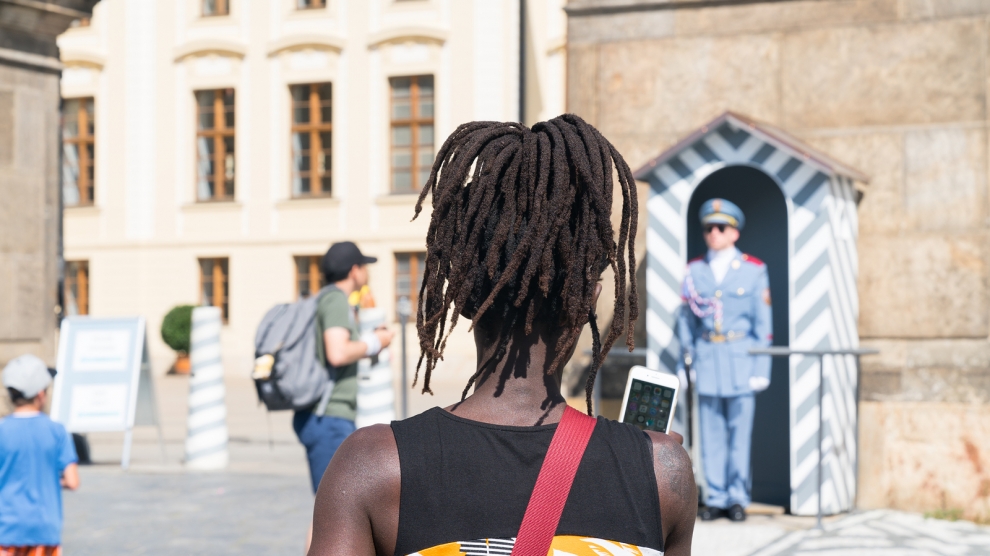 PRAGUE - AUGUST 29: Black woman in selective focus from behind with dreadlocks lifts mobile to yake photo of Guards Prague Castle Guard in blue uniform at attention while other tourists walk around looking at Prague Castle and the guards